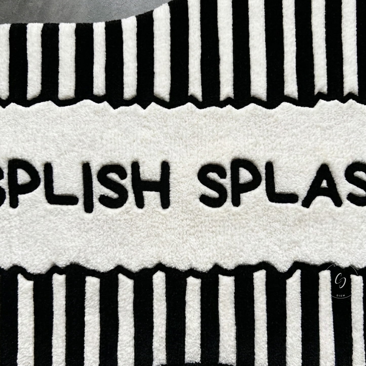 A close-up of a plush hand tufted bathroom entrance rug with bold black vertical striped borders at the top and bottom and the partial text 'SPLISH SPLASH' across the centre.