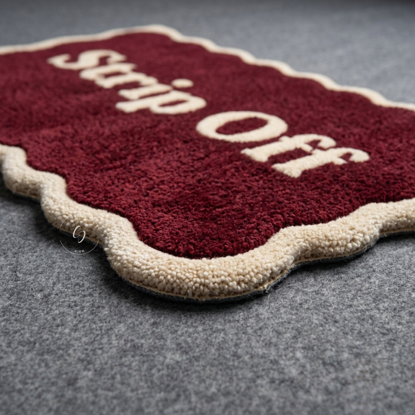 A close-up of a plush hand tufted bathroom entrance rug with a deep burgundy red centre and cream scalloped border, featuring the words 'Strip Off' in cream lettering, placed on a grey textured surface.