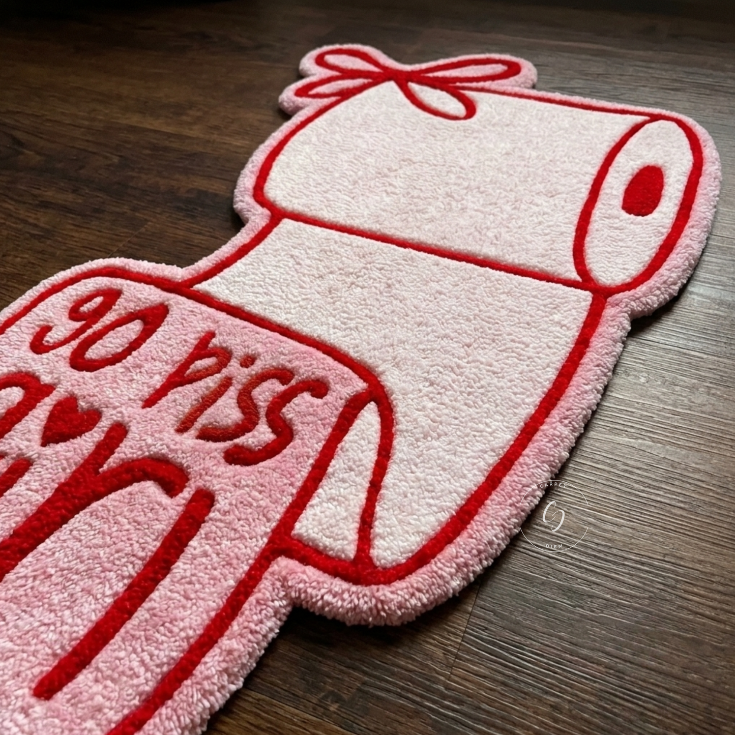 A novelty plush hand tufted bathroom entrance rug shaped like a toilet paper roll with a bow, featuring the text 'go piss girl' in red on a pink and white background, placed on a dark wood floor.