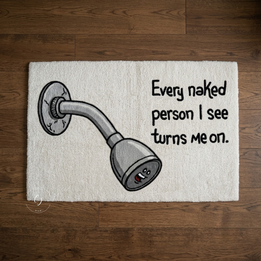A rectangular white hand tufted rug laid flat on a dark wood floor, featuring a cartoon shower head illustration and the text 'Every naked person I see turns me on.'.