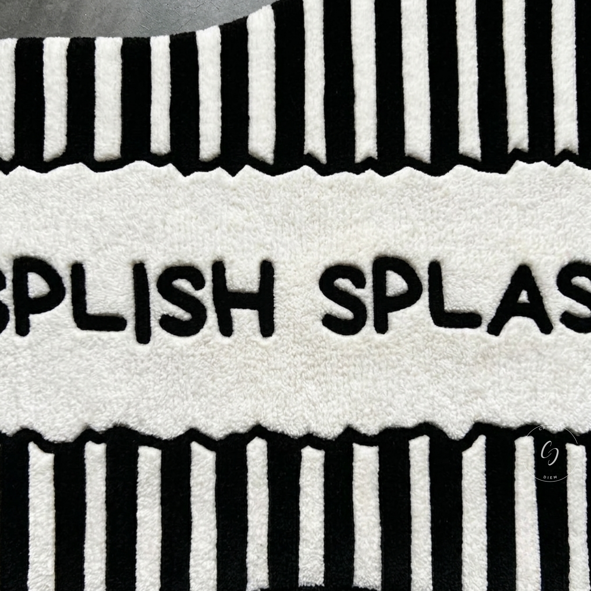 A close-up of a plush hand tufted bathroom entrance rug with bold black vertical striped borders at the top and bottom and the partial text 'SPLISH SPLASH' across the centre.