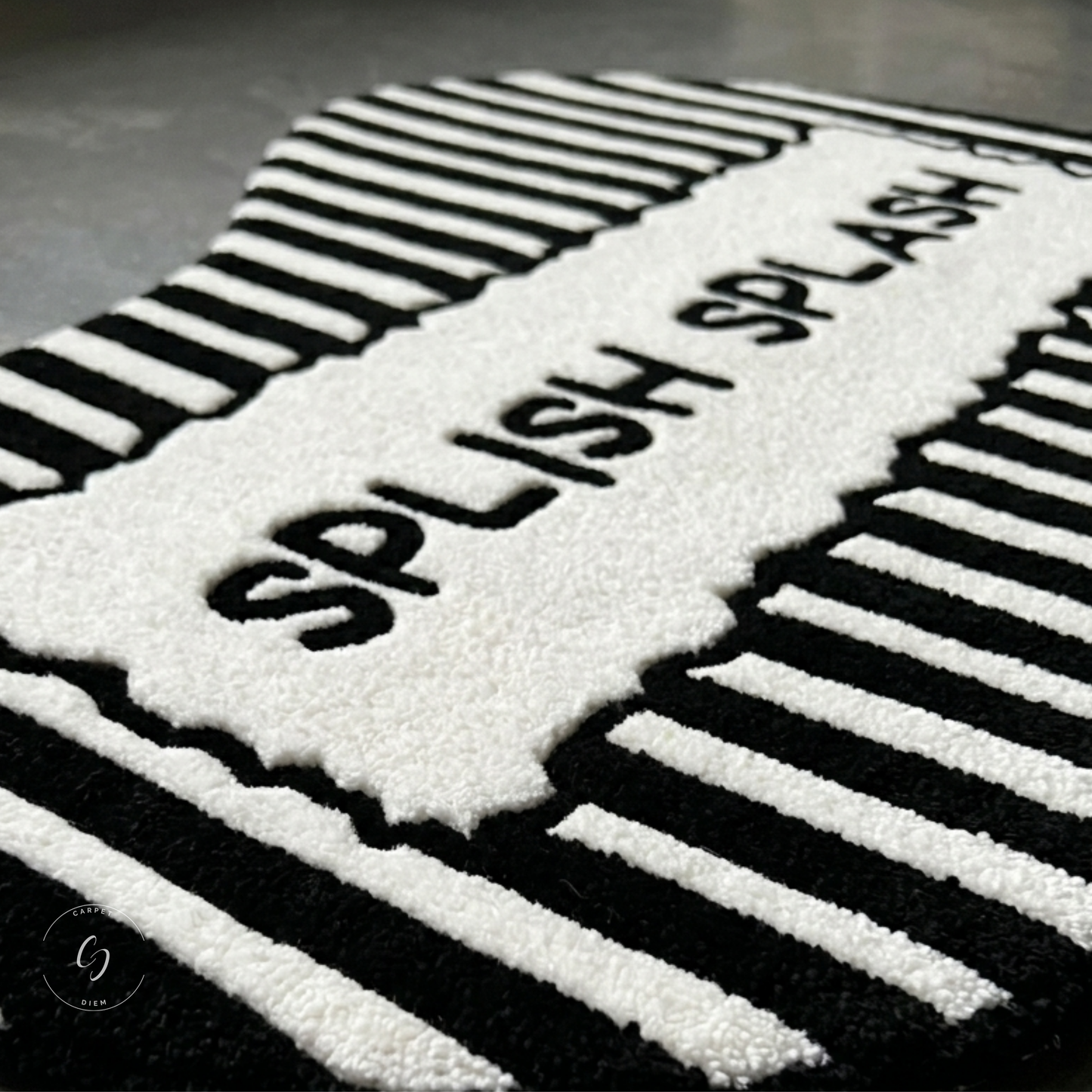 A close-up of a black and white textured hand tufted bathroom entrance rug featuring the words 'SPLISH SPLASH' in raised black letters on a white section, surrounded by bold black and white striped patterns.