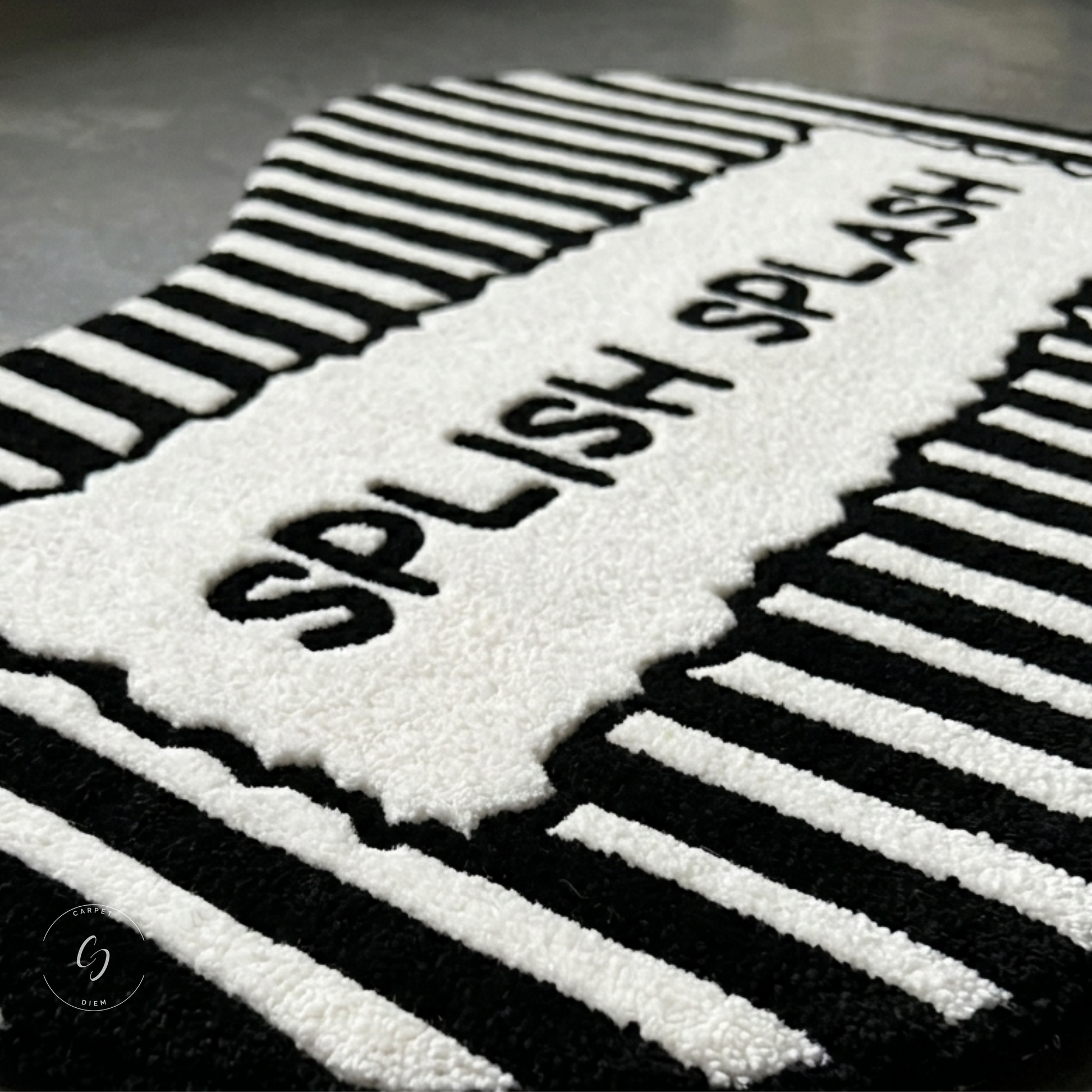 A close-up of a black and white textured hand tufted bathroom entrance rug featuring the words 'SPLISH SPLASH' in raised black letters on a white section, surrounded by bold black and white striped patterns.