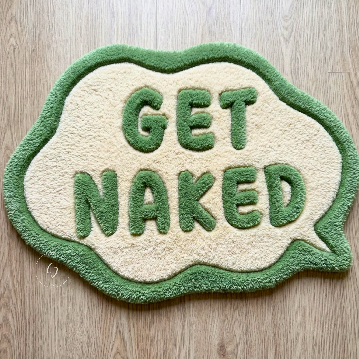 A novelty speech-bubble-shaped hand tufted bathroom entrance rug with the words 'GET NAKED' raised in green on a cream background, placed on a light wood floor.