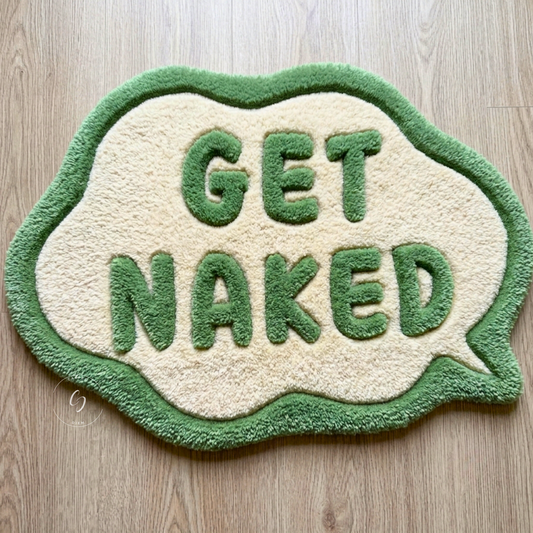 A novelty speech-bubble-shaped hand tufted bathroom entrance rug with the words 'GET NAKED' raised in green on a cream background, placed on a light wood floor.