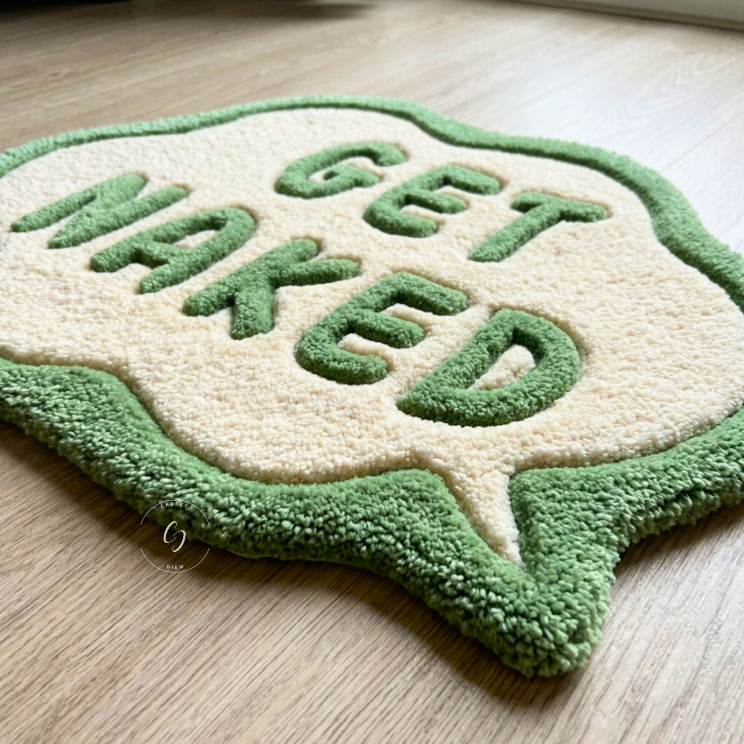 A plush, hand tufted bathroom entrance rug shaped like a speech bubble with the words 'GET NAKED' in green raised letters on a cream background, styled on a light wood floor.