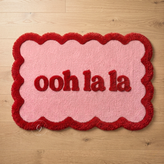 A plush, wavy-edged hand tufted bathroom entrance rug in pink and red with the text 'ooh la la' in red, shown on a light wood floor.