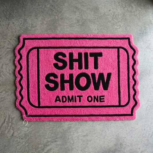 A bright pink hand tufted bathroom entrance rug designed like an old-fashioned ticket, featuring the words 'SHIT SHOW ADMIT ONE' in bold black lettering.