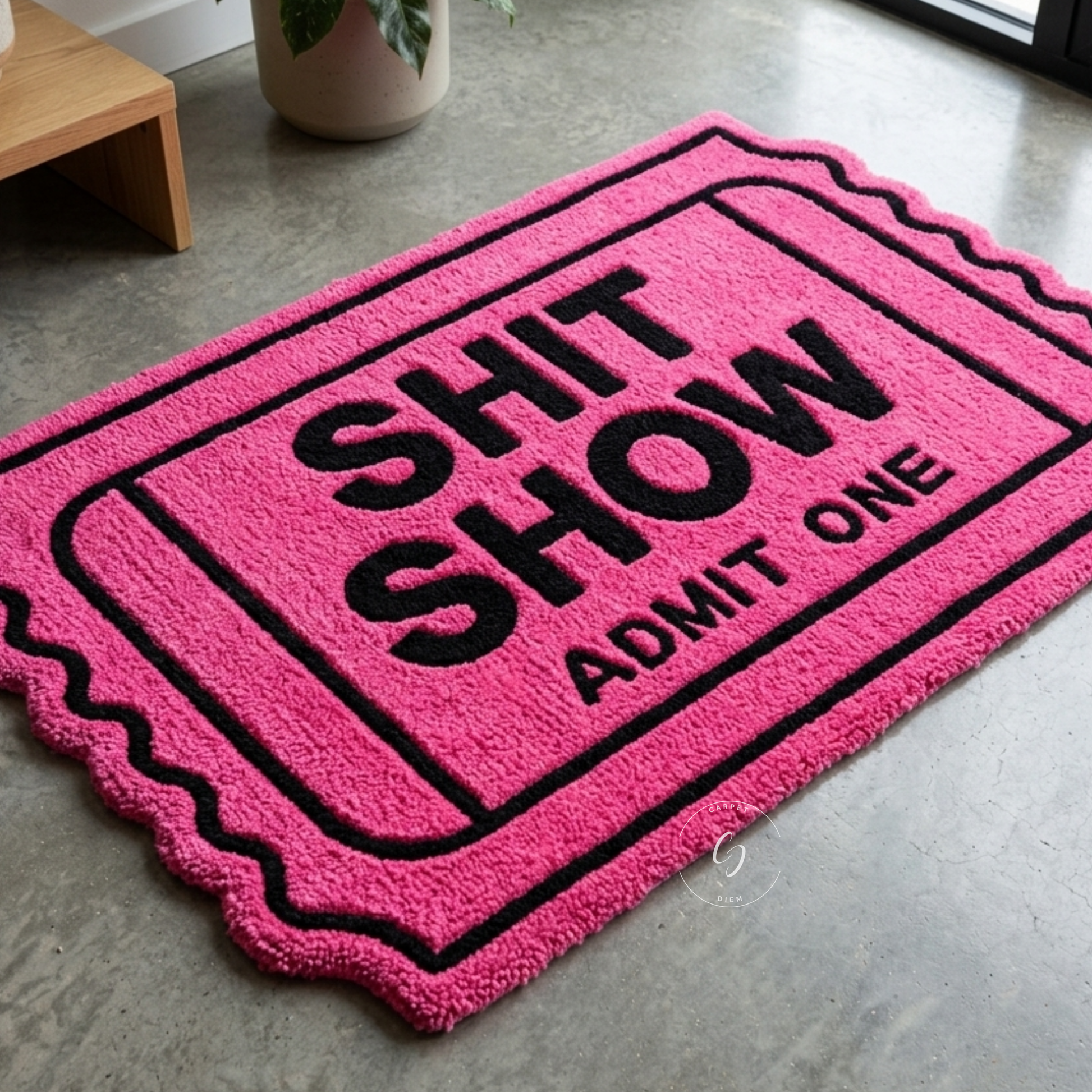 A bright pink rectangular hand tufted bathroom entrance rug with black text reading 'SHIT SHOW ADMIT ONE' in a ticket-style border, photographed on a grey concrete floor indoors.