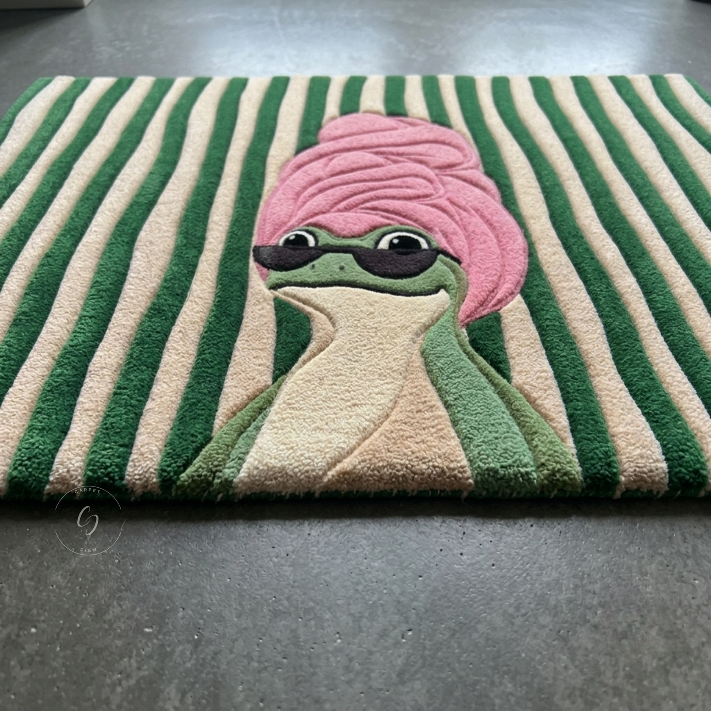 A rectangular hand tufted bathroom entrance rug with green and beige vertical stripes, featuring a raised cartoon frog wearing a pink towel and dark sunglasses, photographed on a dark grey floor.