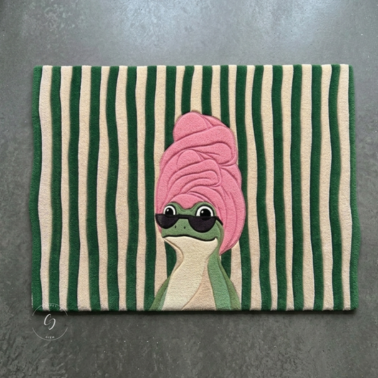 A rectangular hand tufted bathroom entrance rug with vertical green and cream stripes, featuring a cute cartoon frog wearing a pink towel and black sunglasses.