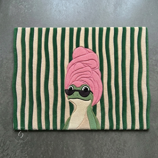 A rectangular hand tufted bathroom entrance rug with vertical green and cream stripes, featuring a cute cartoon frog wearing a pink towel and black sunglasses.