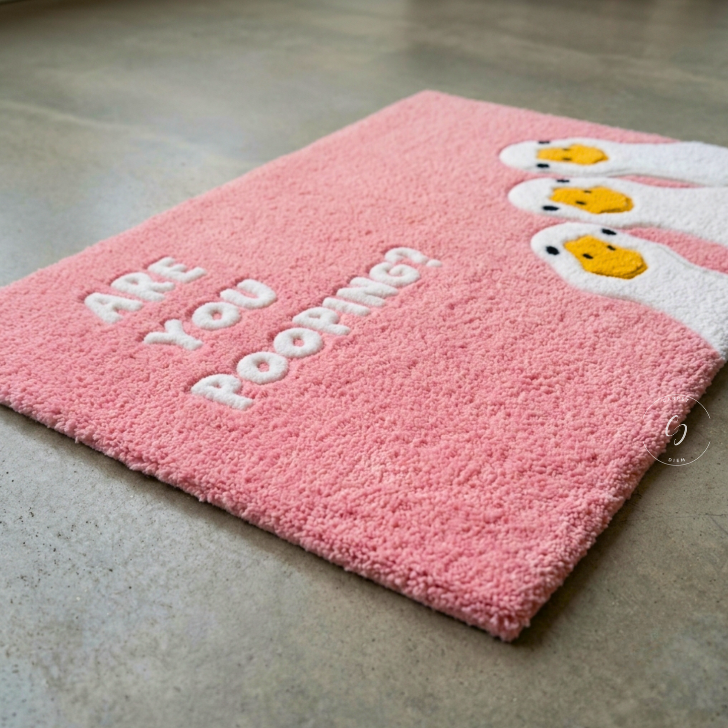 A pink textured hand tufted bathroom entrance rug with white text reading 'ARE YOU POOPING?' and cartoon poop emoji faces on the right, photographed on a grey concrete floor.