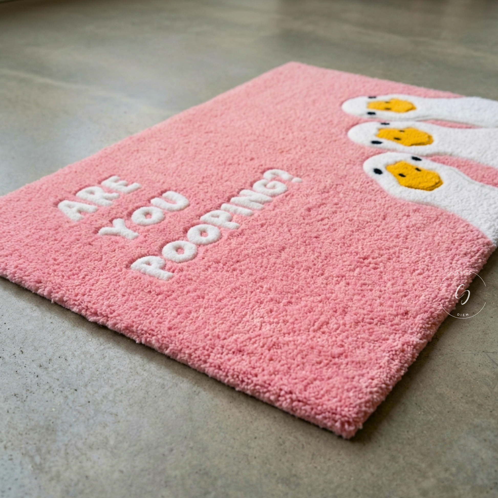 A pink textured hand tufted bathroom entrance rug with white text reading 'ARE YOU POOPING?' and cartoon poop emoji faces on the right, photographed on a grey concrete floor.