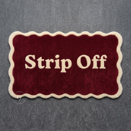 A rectangular plush hand tufted bathroom entrance rug with scalloped cream edges and the words 'Strip Off' in cream lettering on a deep burgundy background.