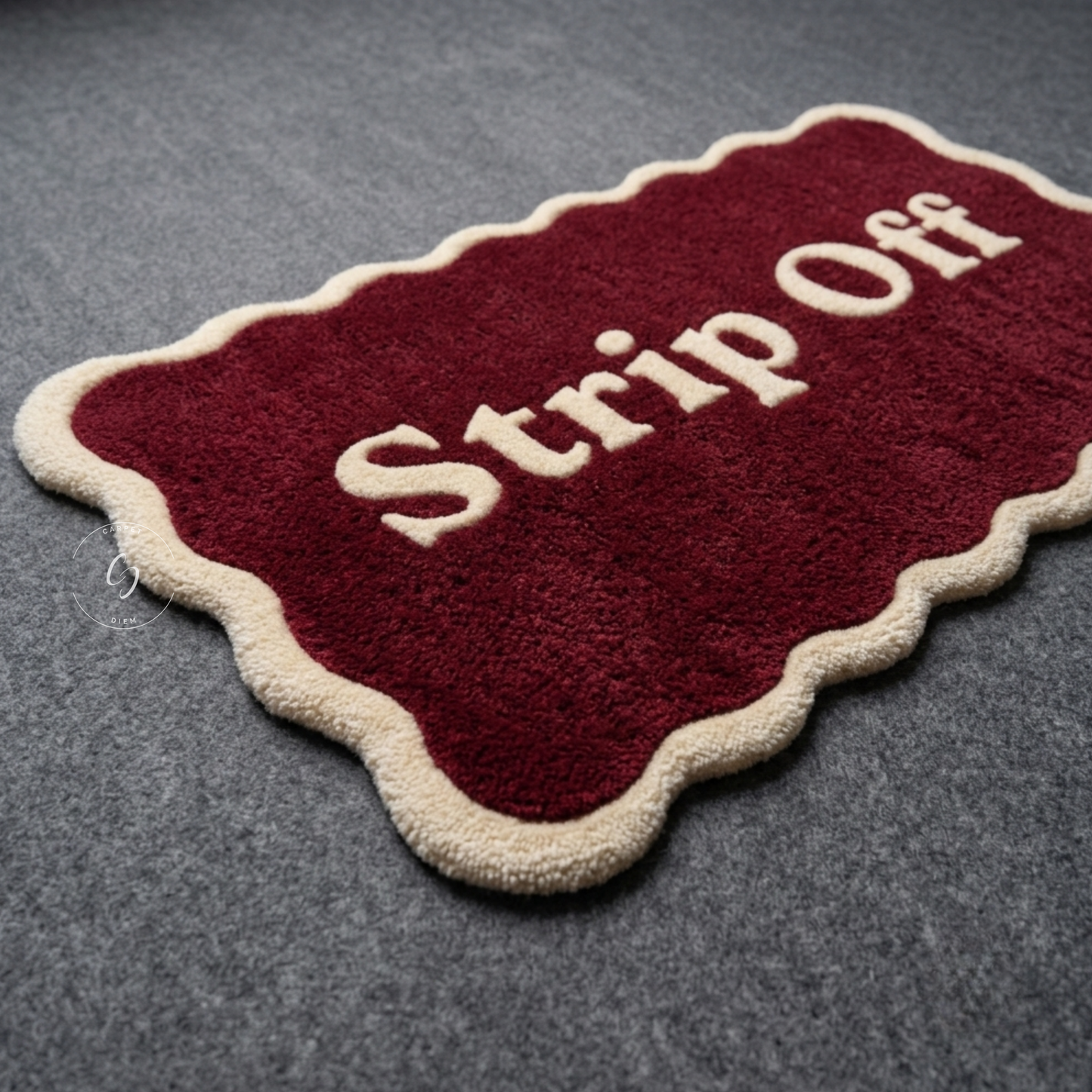 A rectangular red hand tufted bathroom entrance rug with scalloped cream edges and the words 'Strip Off' in cream lettering, placed on a grey textured surface.