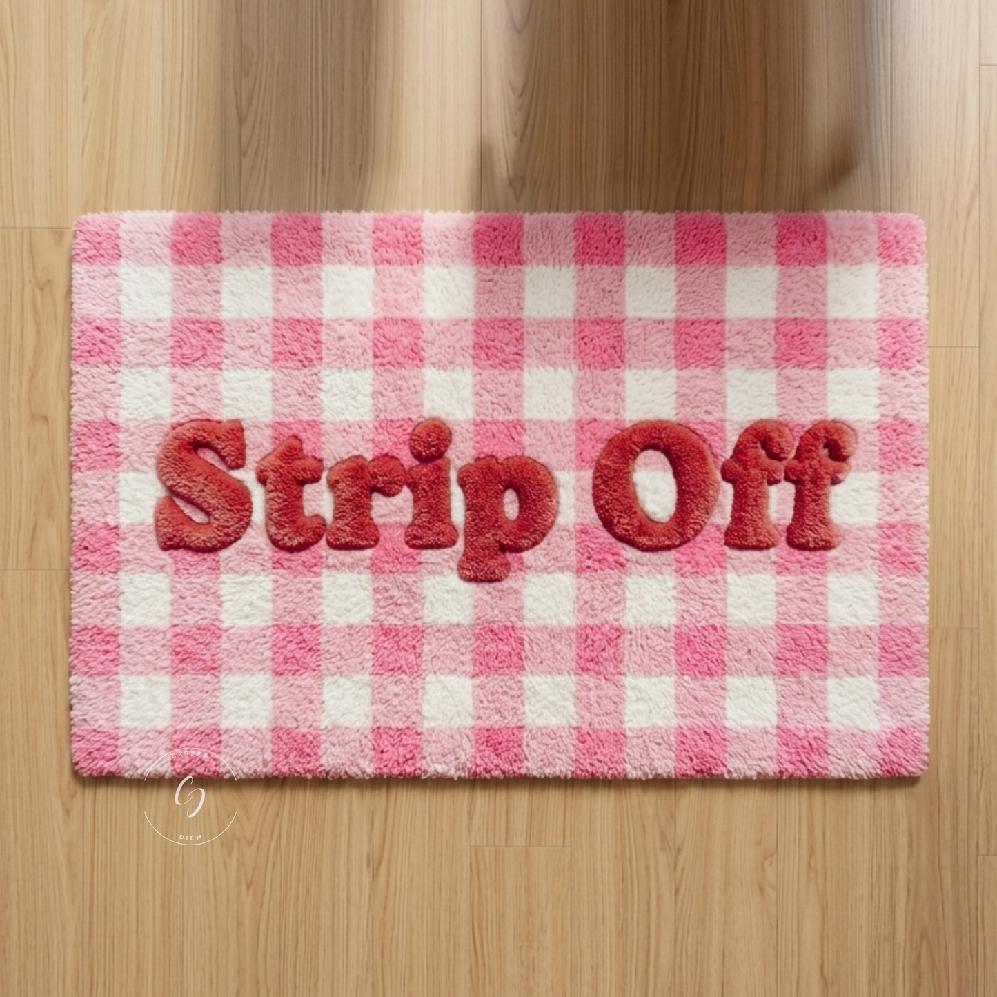 A rectangular pink and white checks hand tufted bathroom entrance rug with the words 'Strip Off' raised in red letters across the centre, shown on a light wood floor.