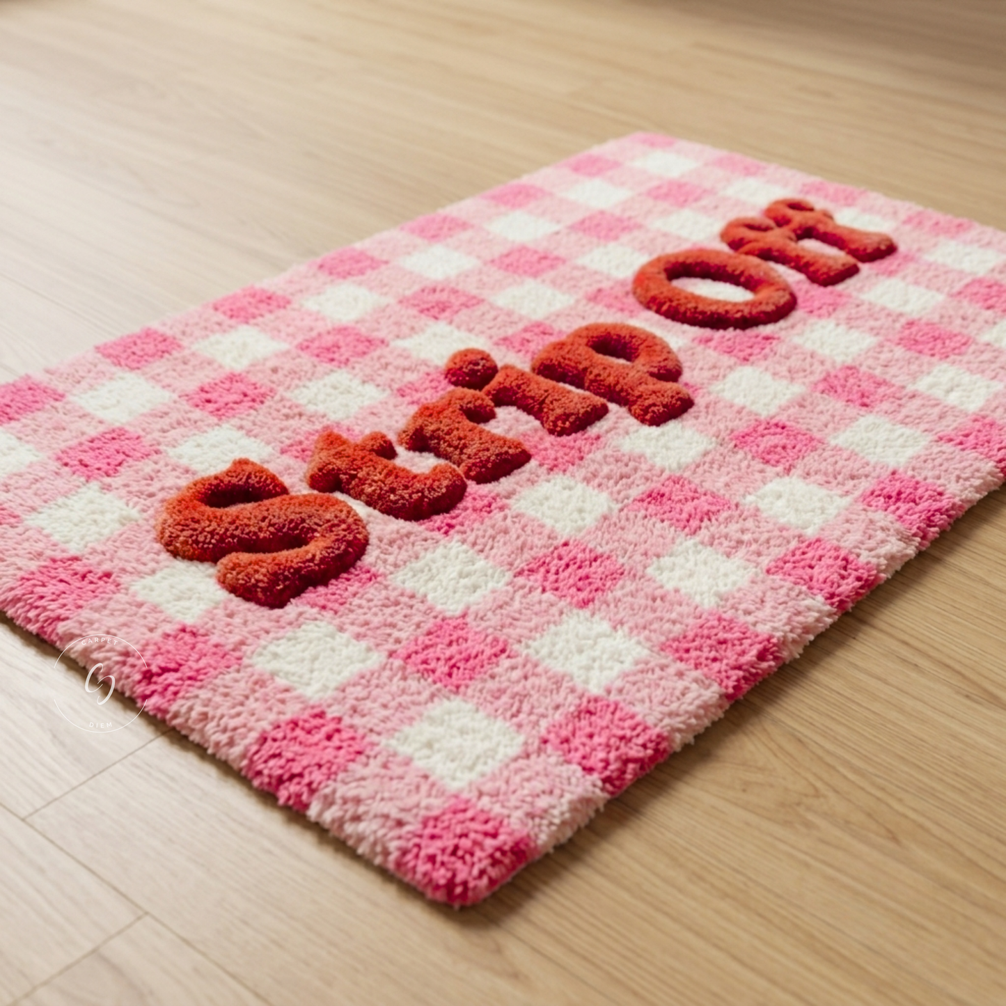 A pink and white checkered plush hand tufted bathroom entrance rug with raised red letters spelling 'STRIP ON' on a light wood floor.