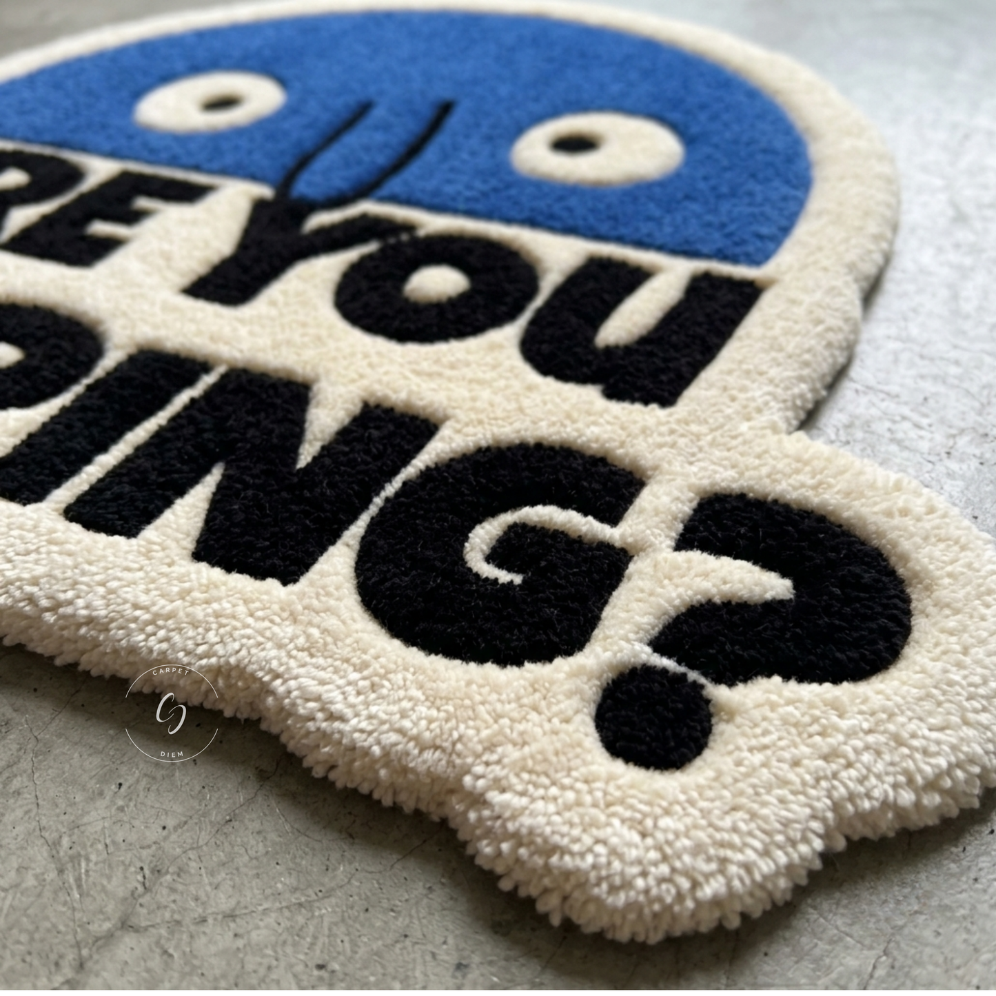 A close-up of a plush, hand tufted bathroom entrance rug featuring a blue cartoon face and the phrase 'ARE YOU POOPING?' in bold black letters on an off-white background, set on a grey concrete floor.