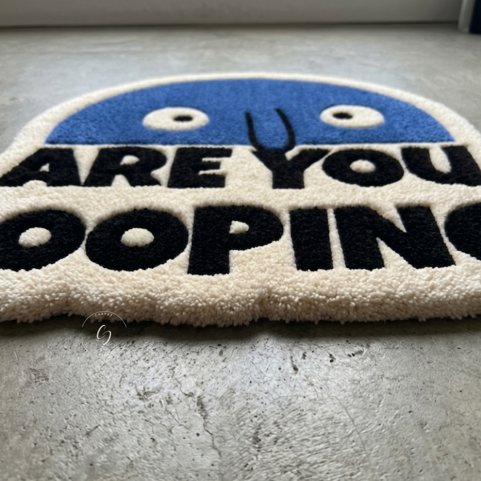A close-up of a textured, hand tufted bathroom entrance rug with a blue smiling face and the text 'ARE YOU pOOPING' in bold black letters on an off-white background, placed on a grey concrete floor.