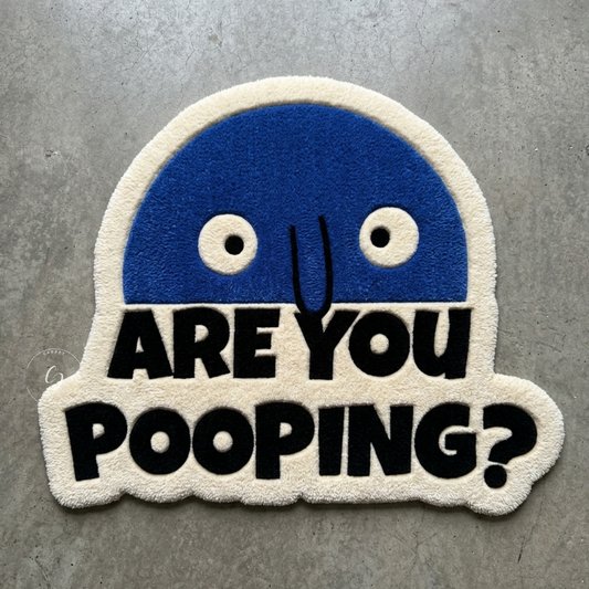 A novelty hand tufted bathroom entrance rug featuring a blue cartoon face above the bold black text 'ARE YOU POOPING?' on a cream fuzzy background, placed on a gray concrete floor.