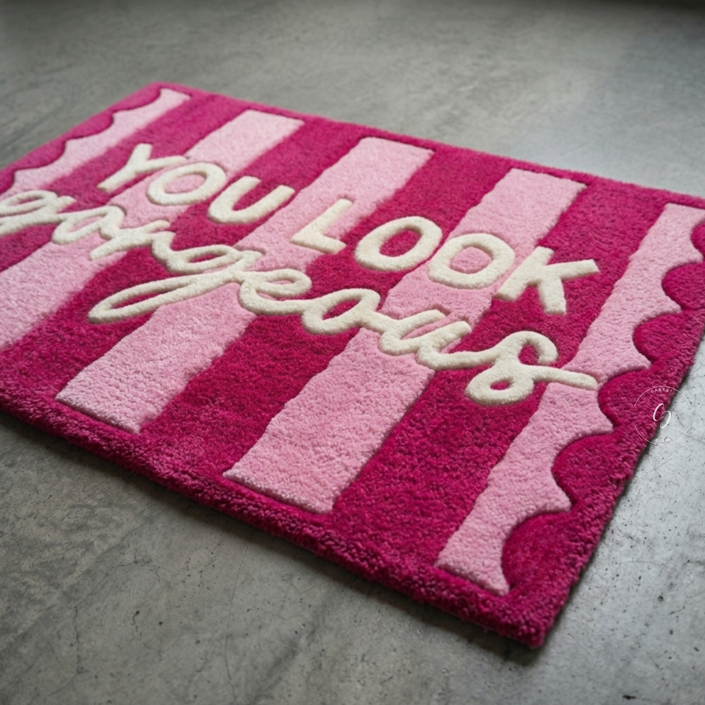 A rectangular pink and magenta striped hand tufted bathroom entrance rug with scalloped edges and the phrase 'YOU LOOK gorgeous' in raised cream lettering, photographed on a grey concrete floor.