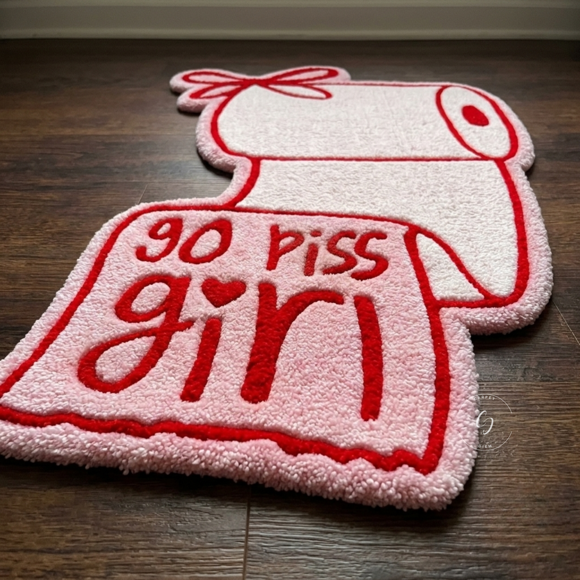A novelty hand tufted bathroom entrance rug shaped like a toilet paper roll with the phrase 'go piss girl' embroidered in red on a pink and white textured surface, placed on a dark wood floor.