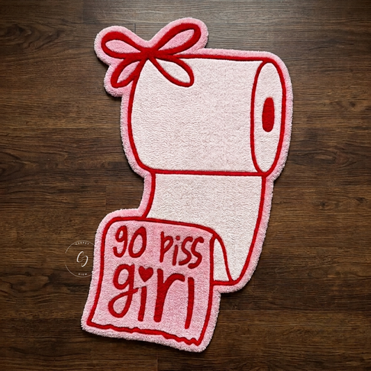 A novelty hand tufted bathroom entrance rug shaped like a toilet paper roll with a bow and a hanging note that reads 'go piss girl' in red text, photographed flat on a dark wood floor.