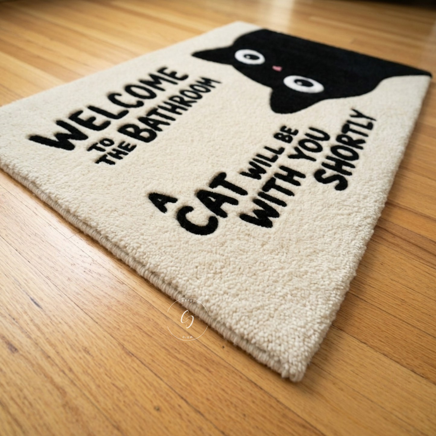 A rectangular beige hand tufted bathroom entrance rug with a black cat graphic and the text 'WELCOME TO THE BATHROOM A CAT WILL BE WITH YOU SHORTLY', photographed on a wooden floor.