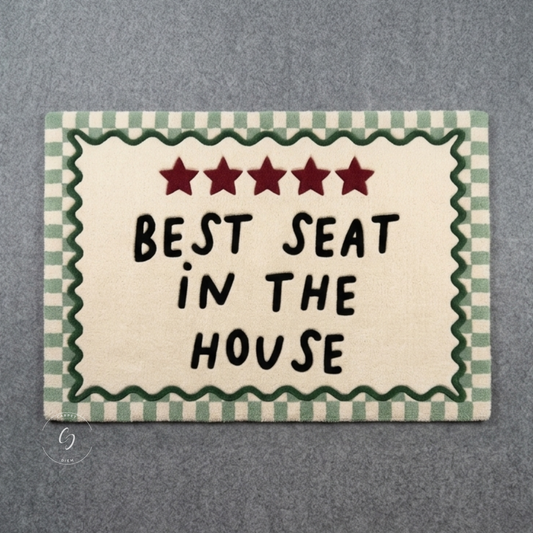A rectangular hand tufted bathroom entrance rug with the phrase “BEST SEAT IN THE HOUSE” in black lettering, five red stars at the top, and a green-and-cream gingham border, shown on a grey textured background.